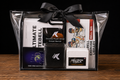 A black gift basket with a black ribbon bow, containing a curated fitness and kettlebell equipment bundle. The bundle includes Ultimate Tebell kettlebell books, a Double Plush Wrist Sweat Band, a black wristband with a white 'K' logo, a Neu Iron Chalk book, and a Bell 365 Exercise Library book, all neatly arranged inside a clear plastic gift bag on a wooden surface against a black background.